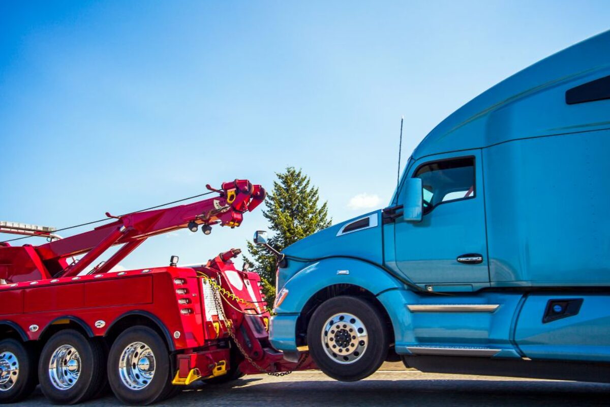 A red tow truck is towing a large blue semi-truck under a clear blue sky, with trees and a building partially visible in the background.